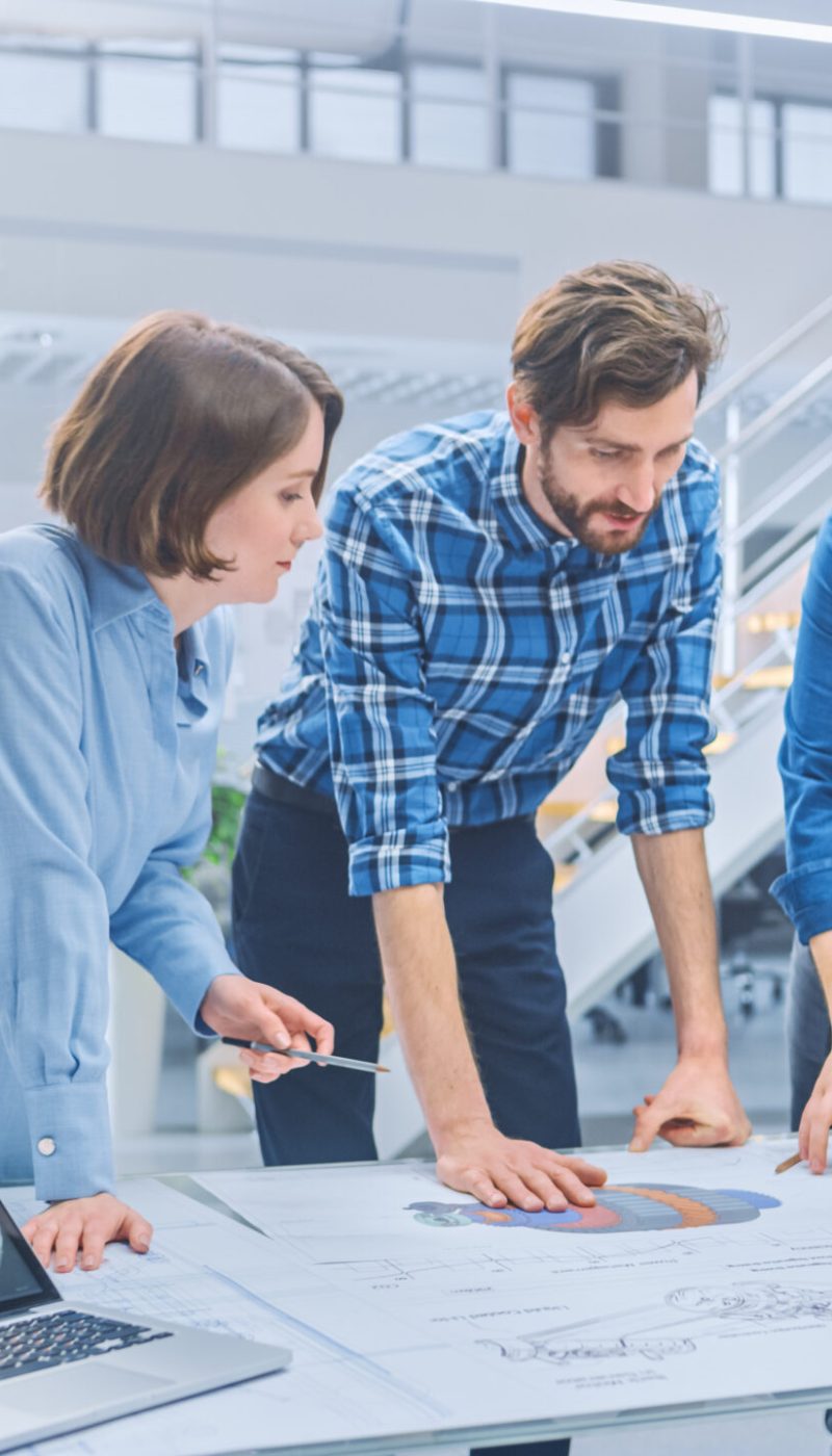 In the Industrial Engineering Facility: Diverse Group of Enginee In the Industrial Engineering Facility: Diverse Group of Engineers and Technicians on a Meeting Gather Around Table with Engine Design Technical Drafts, Have Discussion, Analyse Technology